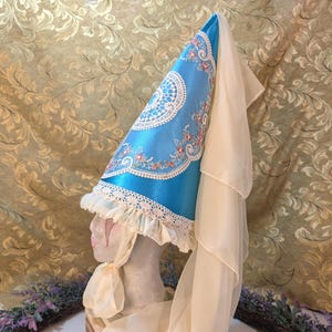 May include: A medieval-style hat with a tall, conical shape. The hat is made of bright blue fabric, adorned with white lace trim and delicate floral embroidery in pink and red. A long, sheer white veil cascades down the back.