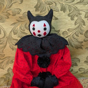 May include: A handmade doll featuring a white face with red diamond markings and a black horned hood. It wears a black eyelet lace collar, black pom-poms, and black gloves over a long-sleeved red dress with a subtle dark red polka dot pattern.