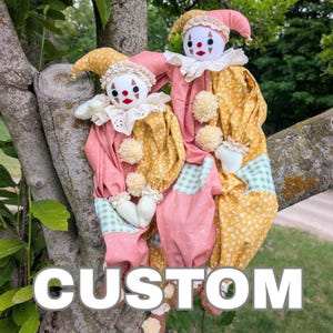 May include: Two fabric clown dolls featuring white faces with painted red and black features, wearing pointed hats with small bells, ruffled collars, and loose-fitting outfits in mustard yellow floral, pink, and green gingham fabric with pom-poms.