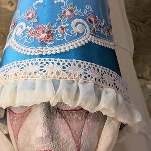May include: A blue and white costume hat with floral embroidery and lace trim. The hat has a ruffled edge and is worn by a painted mannequin head. The hat's design features a combination of textures and colors.