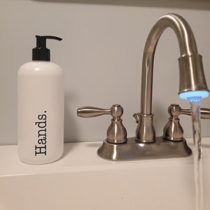 May include: A white soap dispenser with a black pump and the word "Hands." printed vertically in black. The dispenser is next to a silver faucet with water flowing from the spout.