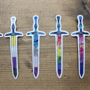 May include: Six colourful sword stickers with different pride flag designs. The stickers are on a wooden surface.