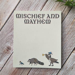 May include: A rectangular notepad with a cream-colored background featuring the text "MISCHIEF AND MAYHEM" in dark brown letters. At the bottom, there is a small blue bird, a raccoon wearing a sword, and a goose wearing a blue jester hat.