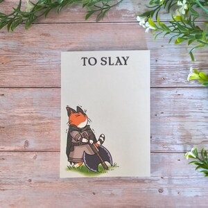 Peut inclure: Un bloc-notes blanc avec le texte "TO SLAY" en haut. Le bloc-notes présente une illustration de dessin animé d'un chat roux et blanc vêtu d'une tenue de guerrier, tenant une grande hache.