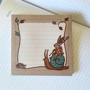 May include: A brown paper notepad with a decorative border featuring a snail and a rabbit wearing armour. The snail is green and brown and the rabbit is brown with a gold and blue patterned armour. The notepad has lined paper and a decorative border with leaves and mushrooms.