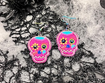 May include: A pair of pink sugar skull earrings with black and white embroidery and colorful accents. The earrings are on a black and white background.