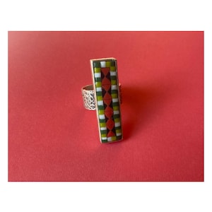 May include: A silver ring with a rectangular mosaic design. The mosaic features a pattern of red, white, green, and yellow tiles. The ring is set against a red background.