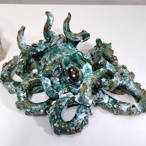 May include: A ceramic octopus sculpture with a textured, glazed finish. The sculpture features a teal and brown color palette with white accents. The tentacles are arranged around a central head, creating a unique decorative piece.