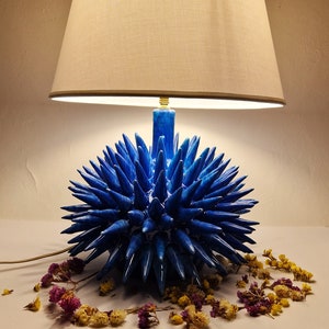 May include: A blue ceramic table lamp with a spiky, sea urchin-like base and a white fabric lampshade.