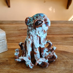 May include: A ceramic octopus sculpture with a brown base, white accents, and blue highlights. The octopus has large eyes and multiple tentacles. The sculpture is placed on a wooden surface.