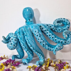May include: A ceramic octopus sculpture with a blue-green glaze. The octopus has eight tentacles with textured details and is posed with its tentacles curled and extended.
