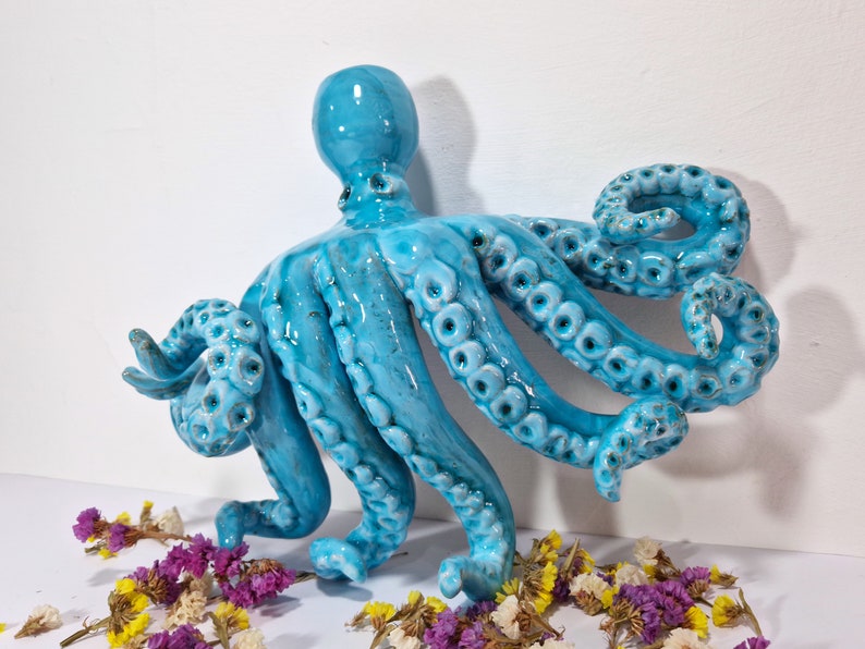 May include: A ceramic octopus sculpture with a blue-green glaze. The octopus has eight tentacles with textured details and is posed with its tentacles curled and extended.