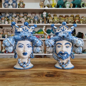 Könnte beinhalten: Zwei Keramikkopfvasen, eine männliche und eine weibliche, mit blauen und weißen Details. Die Vasen haben Kronen, verziertes Haar und Schmuck. Der Hintergrund zeigt Regale mit ähnlichen Keramikkunstwerken.