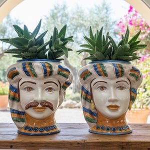 May include: Two ceramic face planters, one with a moustache and the other without, each holding green succulents. The planters have a white base with blue, orange, and green striped accents and are set on a wooden surface.