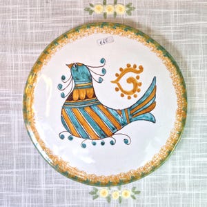 May include: A white ceramic plate with a yellow and green border. The plate features a hand-painted design of a blue and yellow bird with a yellow sun behind it. The price tag on the plate reads "£45".