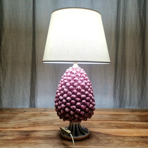 Può includere: Una lampada da tavolo in ceramica rosa a forma di pigna con un paralume bianco. La lampada ha una base bianca e blu.
