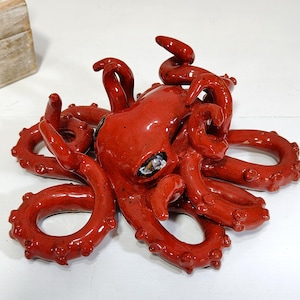 May include: A glossy, red ceramic octopus sculpture with detailed tentacles and a textured surface. The octopus has a detailed head and is a decorative item.