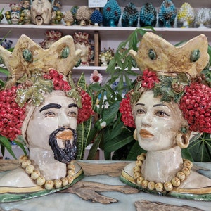 May include: Two ceramic sculptures of human heads with crowns, red berries, and green leaves. The sculptures are painted in shades of white, yellow, green, and red.