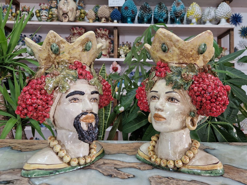 May include: Two ceramic sculptures of human heads with crowns, red berries, and green leaves. The sculptures are painted in shades of white, yellow, green, and red.