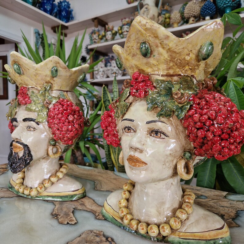 May include: Two ceramic sculptures of human heads with crowns, floral embellishments, and red berries. The sculptures are painted in shades of yellow, green, red, and brown.