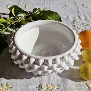 Puede incluir: Tazón de cerámica blanco con un exterior texturizado y puntiagudo. El tazón está vacío y se encuentra sobre un mantel blanco con limones y una naranja a su lado.