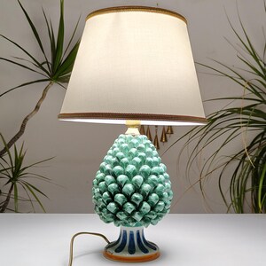 Può includere: Una lampada da tavolo in ceramica con una base a forma di pigna verde e un paralume color crema. La base presenta un design dettagliato e strutturato, e il paralume è rifinito con un bordo decorativo. La lampada è su una superficie bianca.