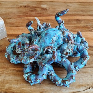 May include: A ceramic octopus sculpture with a textured, weathered appearance. The sculpture features a blend of blue and brown tones, with detailed tentacles and a central body. The artwork is displayed on a wooden surface.