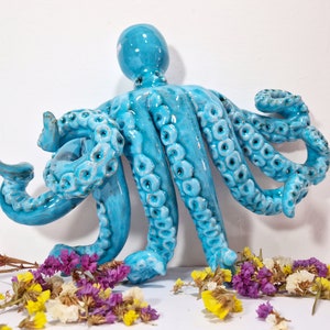 May include: A blue ceramic octopus sculpture with detailed tentacles. The octopus is sitting on a white surface with dried flowers.