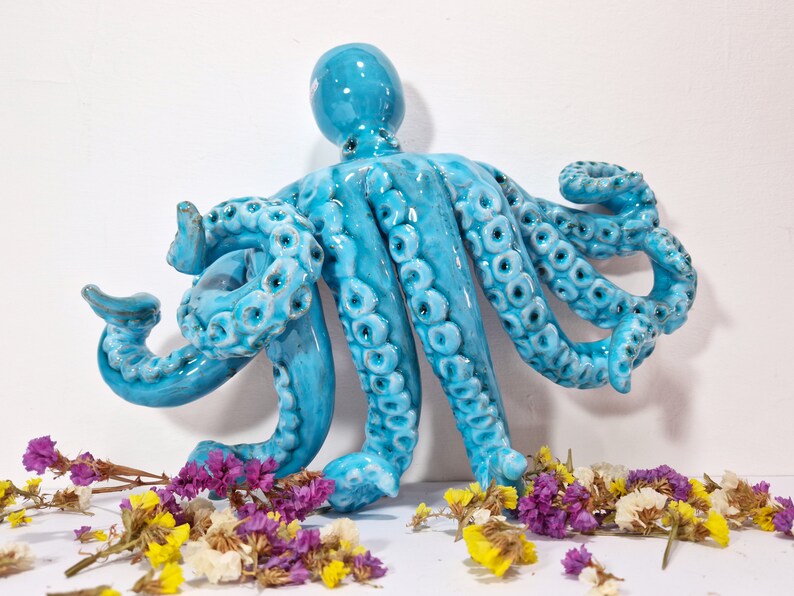 May include: A blue ceramic octopus sculpture with detailed tentacles. The octopus is sitting on a white surface with dried flowers.