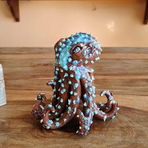 May include: A brown ceramic octopus sculpture with turquoise accents. The octopus has a textured surface with small, raised bumps. The sculpture is on a wooden surface, showcasing a handcrafted nautical theme.
