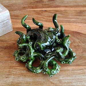 May include: A dark green ceramic octopus sculpture with multiple tentacles. The sculpture has a glossy finish and is placed on a wooden surface. The tentacles are curled and detailed, creating a unique decorative piece.