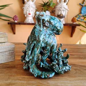 May include: A ceramic octopus sculpture with a turquoise and bronze glaze. The octopus has detailed tentacles and sits on a wooden surface. Decorative ceramic heads and plants are in the background.