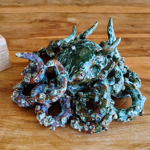 May include: A ceramic sculpture of an octopus with a textured surface. The sculpture features a dark green head and tentacles in shades of blue, brown, and white. The artwork is displayed on a wooden surface.