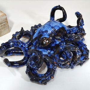 May include: A ceramic octopus sculpture with a glossy, mottled blue and black glaze. The tentacles curl and extend, and the sculpture has a detailed, textured surface. The artwork is a decorative piece.