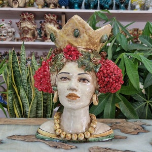 May include: A ceramic bust of a woman with a crown, red berries, and a yellow beaded necklace. The bust is painted in shades of white, yellow, green, and red.