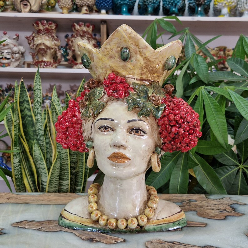 May include: A ceramic bust of a woman with a crown, red berries, and a yellow beaded necklace. The bust is painted in shades of white, yellow, green, and red.