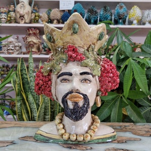 May include: Ceramic sculpture of a king's head with a crown, beard, and a necklace of beads. The sculpture is decorated with red berries and green leaves.