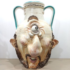 May include: A white ceramic vase shaped like a face with a single large eye, a long nose, and a beard. The vase has two handles and a turquoise blue rim.