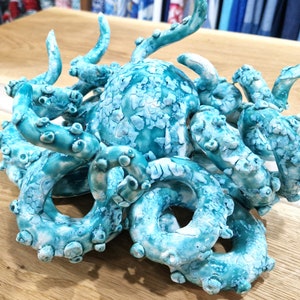 May include: A ceramic octopus sculpture with a teal blue and white glaze. The octopus has eight tentacles that are curled and twisted together.