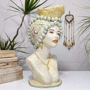 May include: A cream-coloured ceramic bust of a woman. The bust features a detailed headpiece, adorned with light blue spheres and a yellow crown. The woman wears a gold necklace and earrings. The bust is a decorative item.