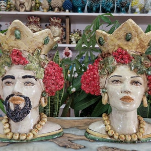 May include: Two ceramic sculptures of human heads with crowns, red berries, and green leaves. The sculptures are painted in shades of white, yellow, green, and red.