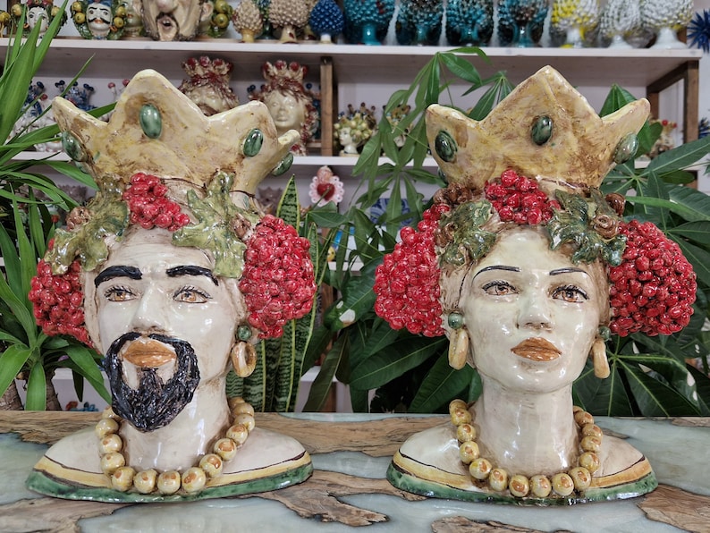 May include: Two ceramic sculptures of human heads with crowns, red berries, and green leaves. The sculptures are painted in shades of white, yellow, green, and red.
