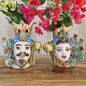May include: Two ceramic vases shaped like faces, one male and one female, with colorful details and floral designs. The vases are decorated with cactus and lemons. The vases are filled with pink flowers.