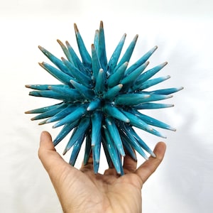 May include: A turquoise ceramic sculpture with a spiky, starburst design. The tips of the spikes are a light brown color. The sculpture is held in a hand against a white background. The object is a decorative art piece.
