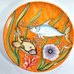 May include: A round orange ceramic plate with a painted design of two fish, seaweed, and a purple sea urchin. The fish are a light blue swordfish and a brown and yellow fish.