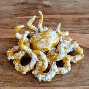 May include: A ceramic octopus sculpture in shades of yellow and white. The octopus has multiple curled tentacles and a rounded head. The sculpture is placed on a wooden surface.