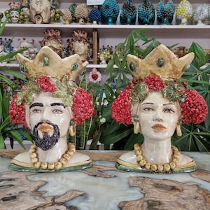 May include: Two ceramic sculptures of human heads with crowns, red berries, and green leaves. The sculptures are painted in shades of yellow, red, green, and brown. They are wearing necklaces of white beads.