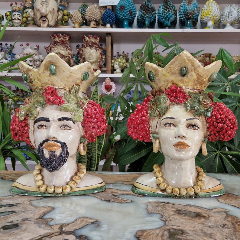 May include: Two ceramic sculptures of human heads with crowns, red berries, and green leaves. The sculptures are painted in shades of yellow, red, green, and brown. They are wearing necklaces of white beads.
