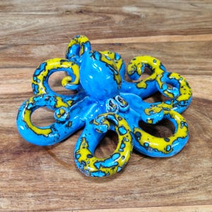 May include: A blue and yellow ceramic octopus figurine with a speckled glaze. The octopus has eight tentacles and is curled up in a ball.