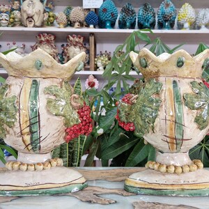 May include: Two ceramic vases with a crown-like top, decorated with green leaves, red berries, and a yellow and green striped pattern. The vases are on a white surface with a green and yellow border. The text "Tradizioni" is visible on the base of one vase.
